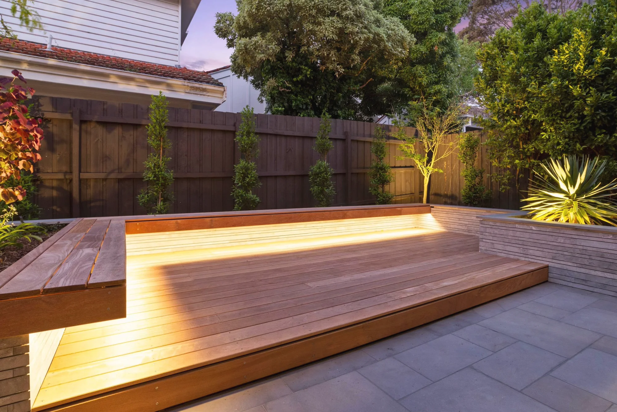 Hardwood deck edged with an LED strip beside stone paving and an uplit yucca at dusk