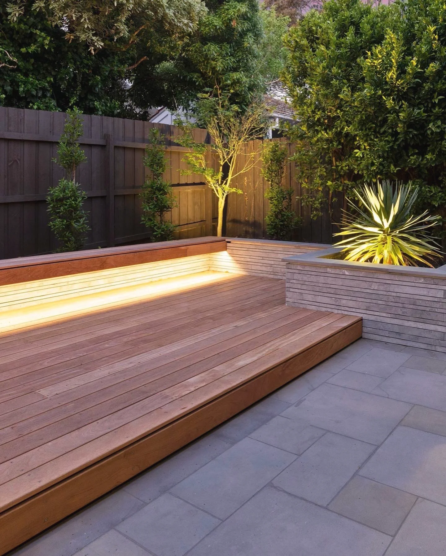 Hardwood deck step with an LED strip, a stone-clad planter and stone paving at dusk