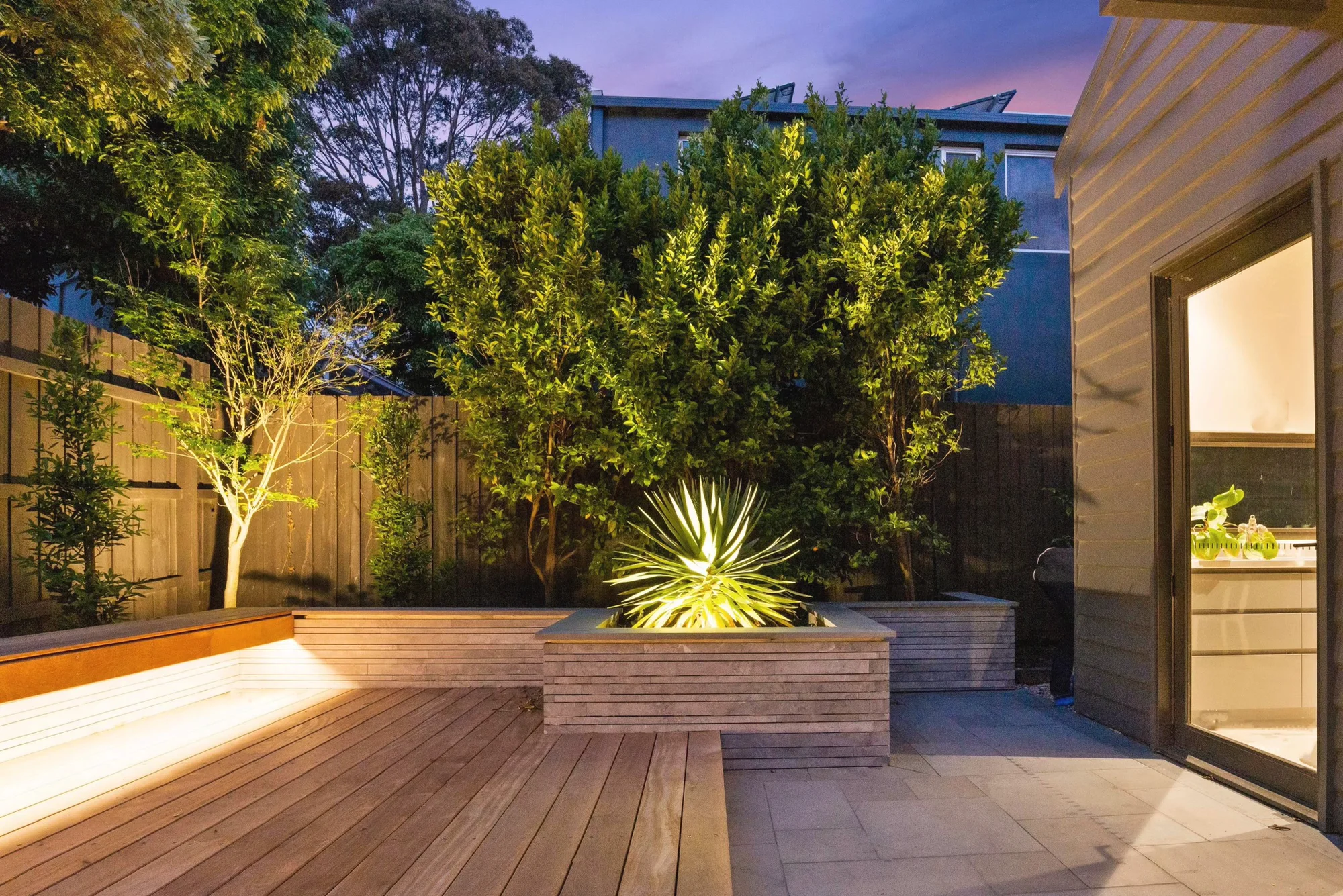 Hardwood deck beside an uplit yucca in a stone-clad planter at dusk