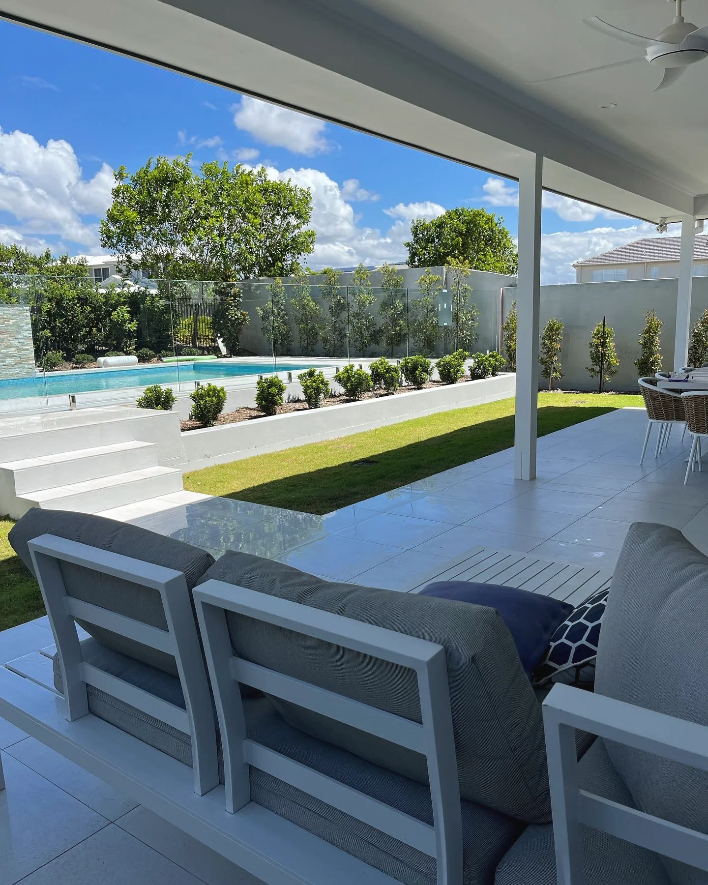 View from a covered patio over rendered steps, a raised planter and lawn to a pool behind glass