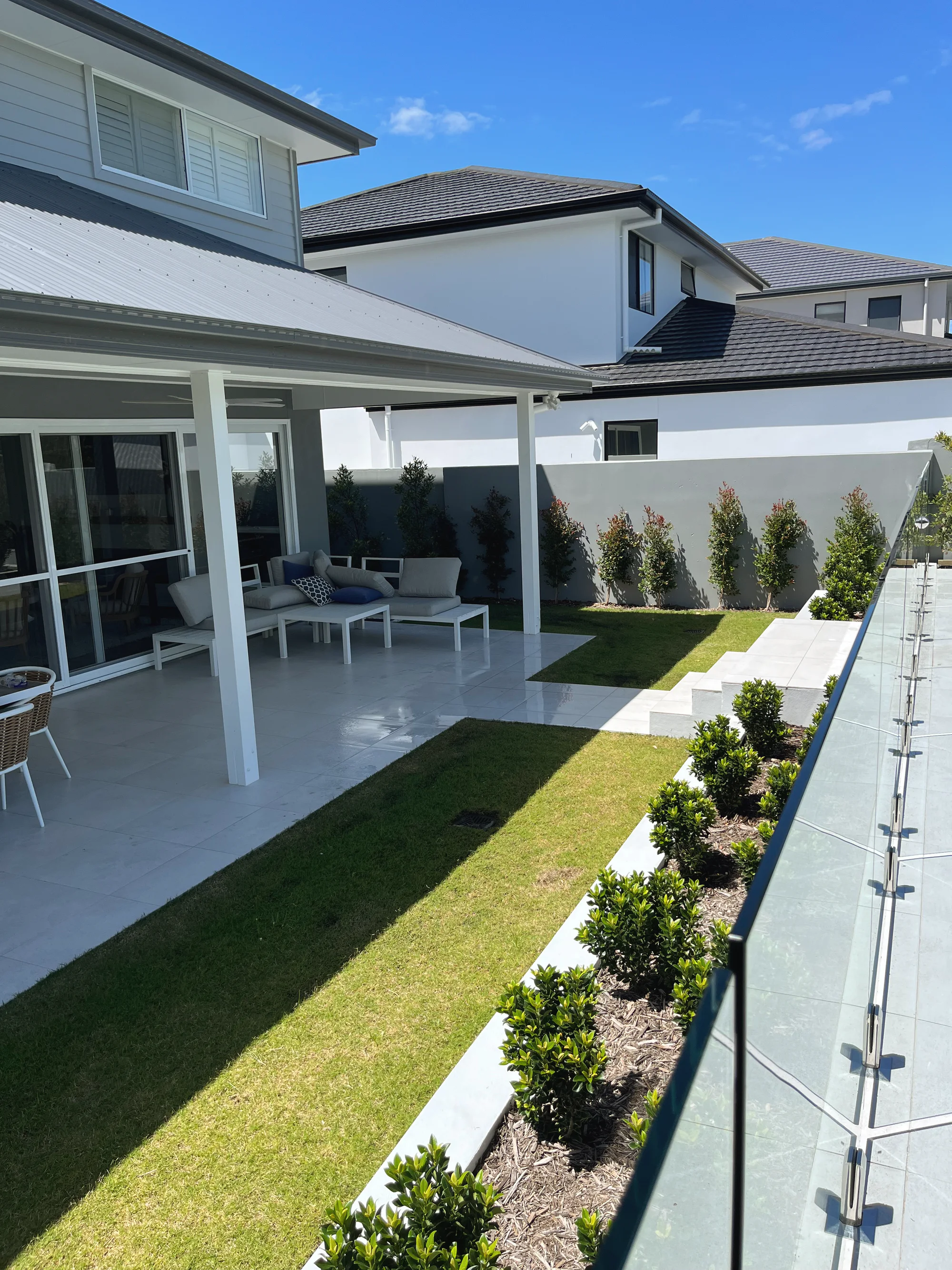 Side lawn between a paved patio and hedges in raised rendered planters along a glass pool fence
