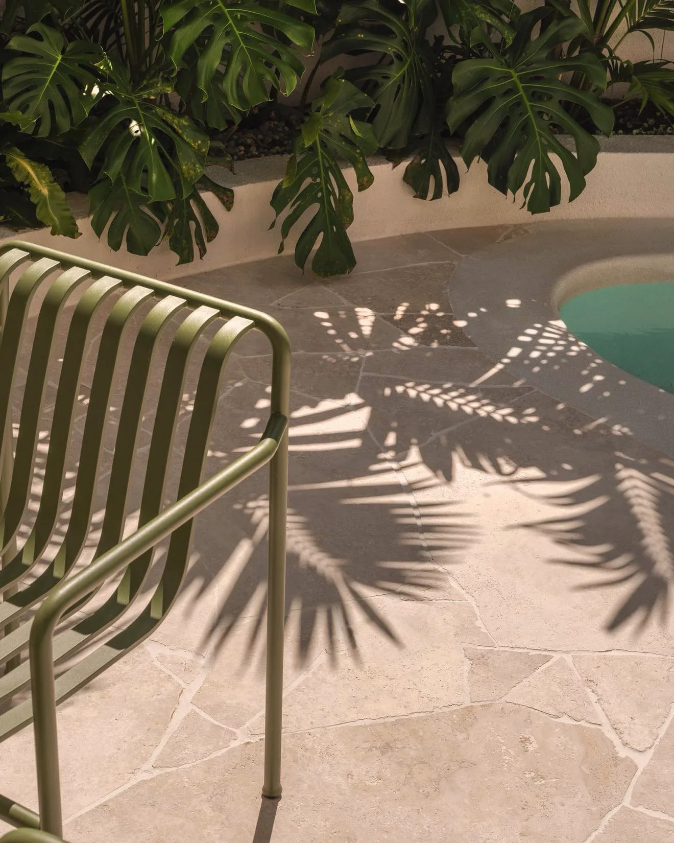 Palm shadows on natural stone crazy paving beside a plunge pool, with a green outdoor chair and monstera in a planter