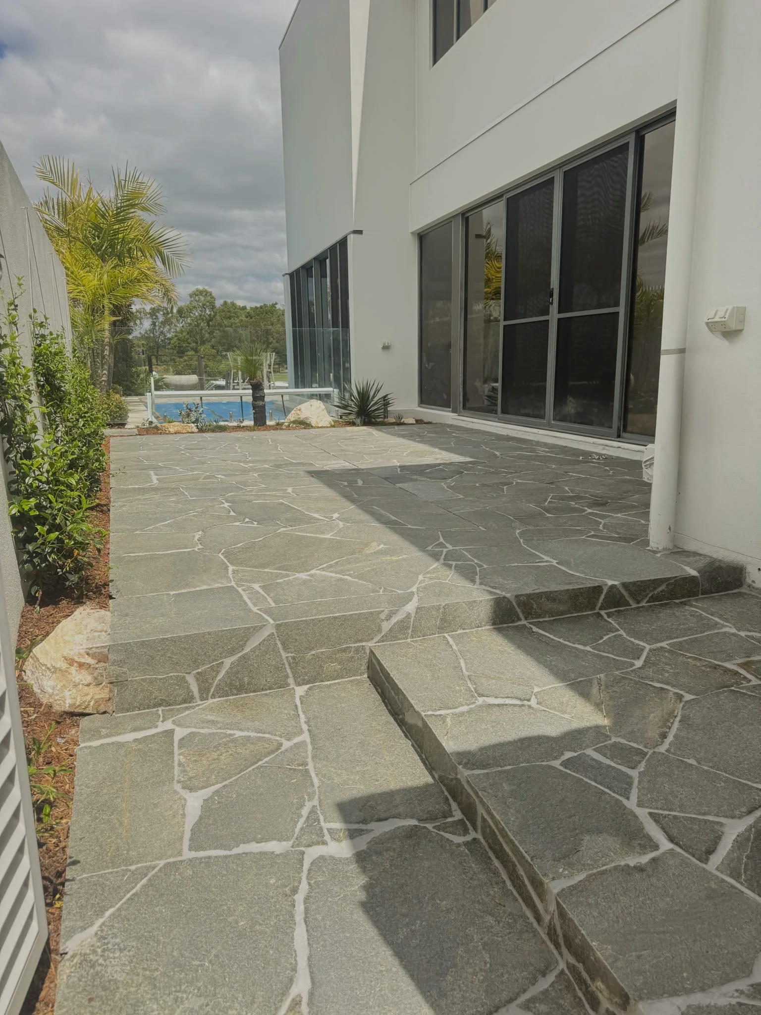 Crazy paved terrace and steps along a white two-storey home, pool beyond