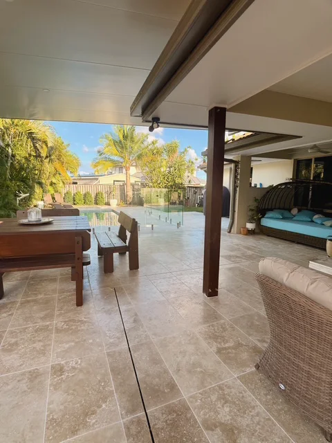Covered alfresco laid in large pale tiles with a slot drain, looking out to a pool behind frameless glass and palms