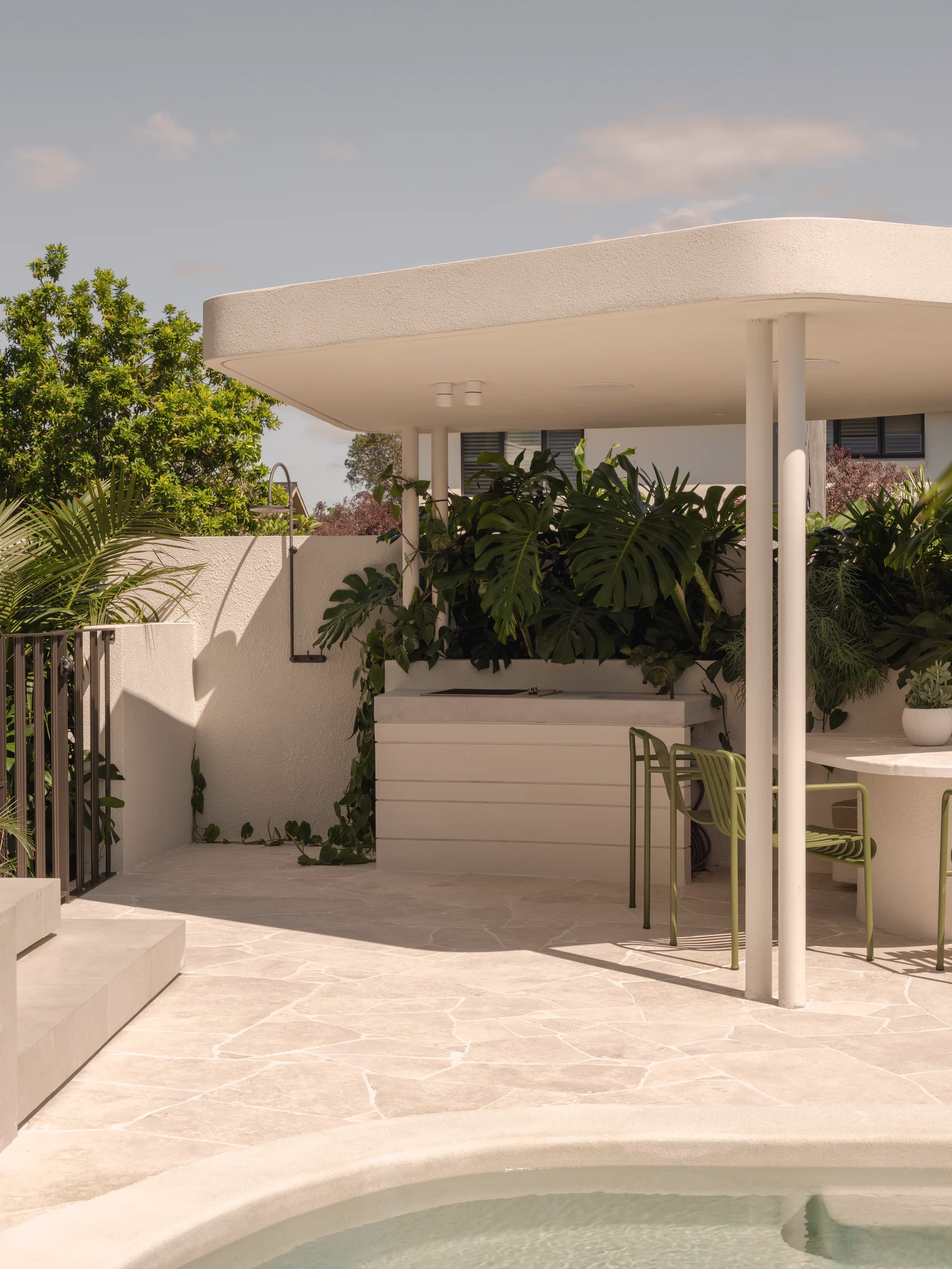 Crazy paved pool terrace with an outdoor kitchen bench, green chairs and monstera planting