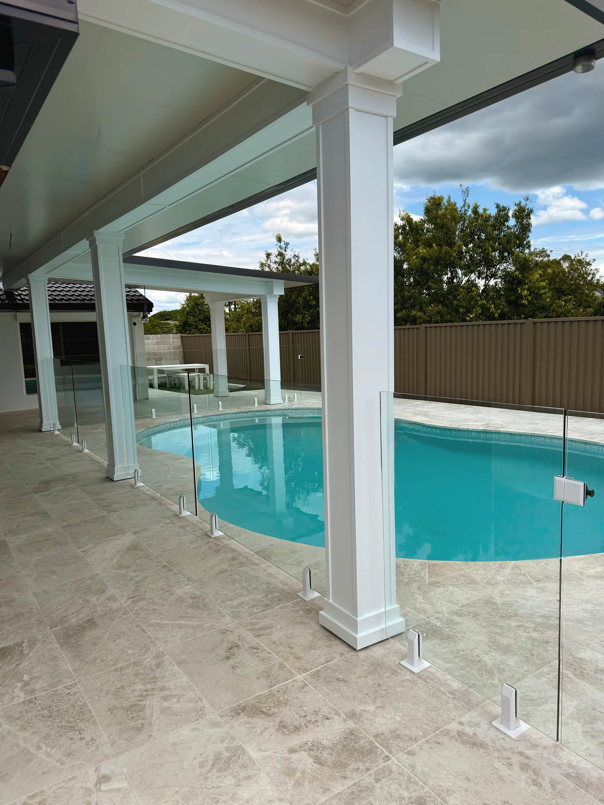 Pale marble-look pavers around a curved pool behind a frameless glass fence
