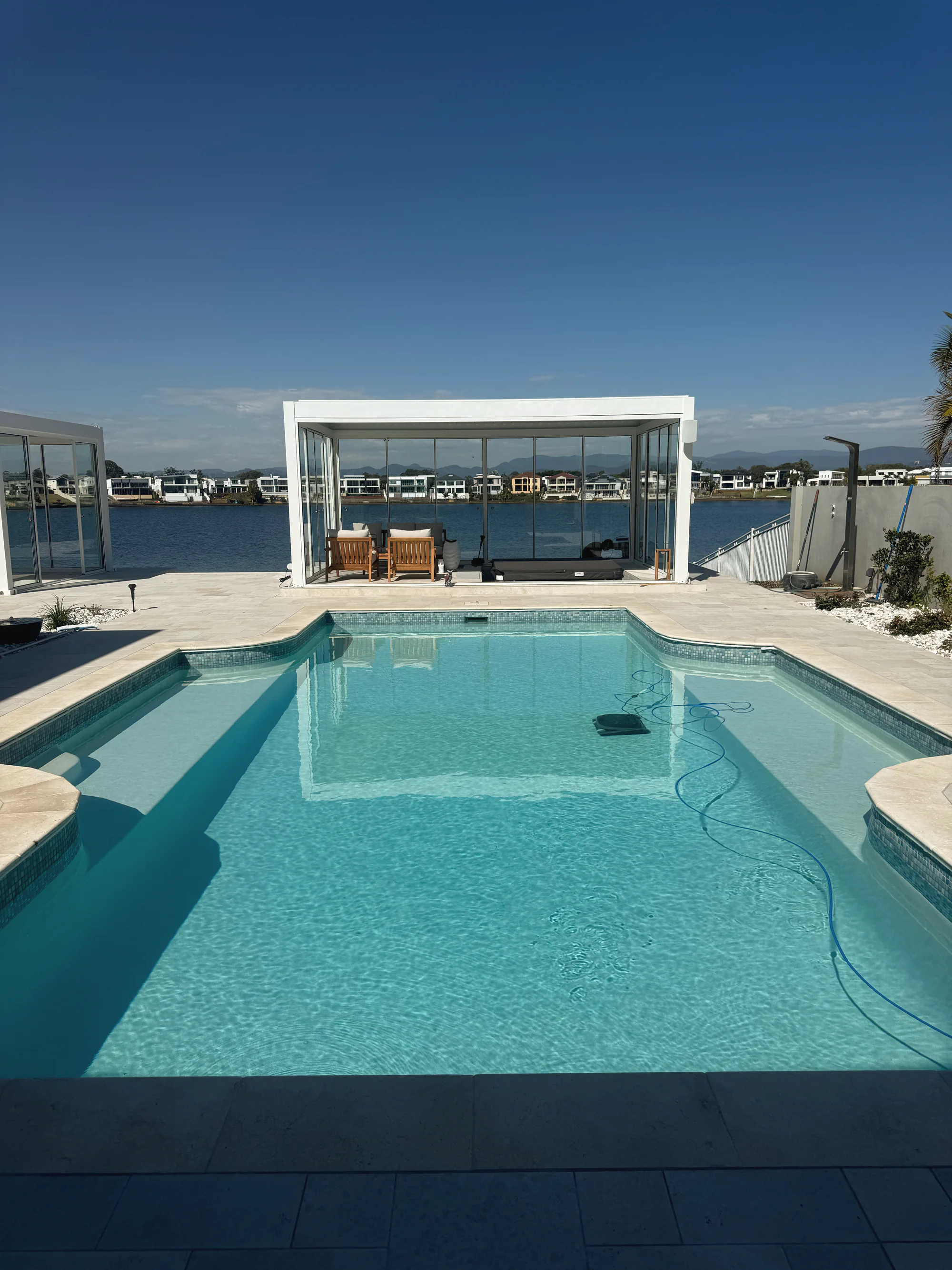 Pool with travertine coping on a waterfront block, a glass pavilion at the far end and the water beyond