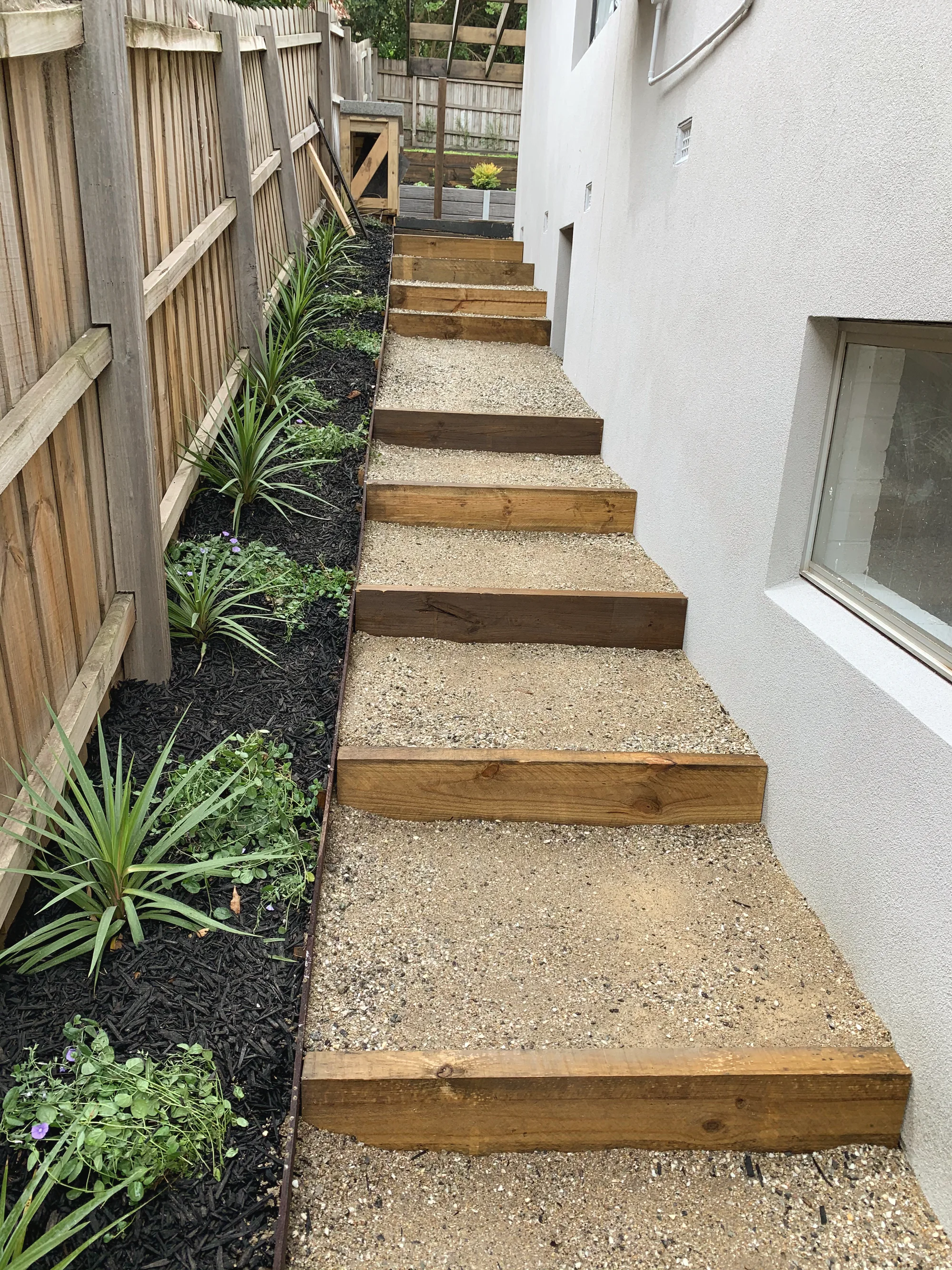 Timber sleeper steps with a gravel fill climbing a narrow side path, planted along the fence