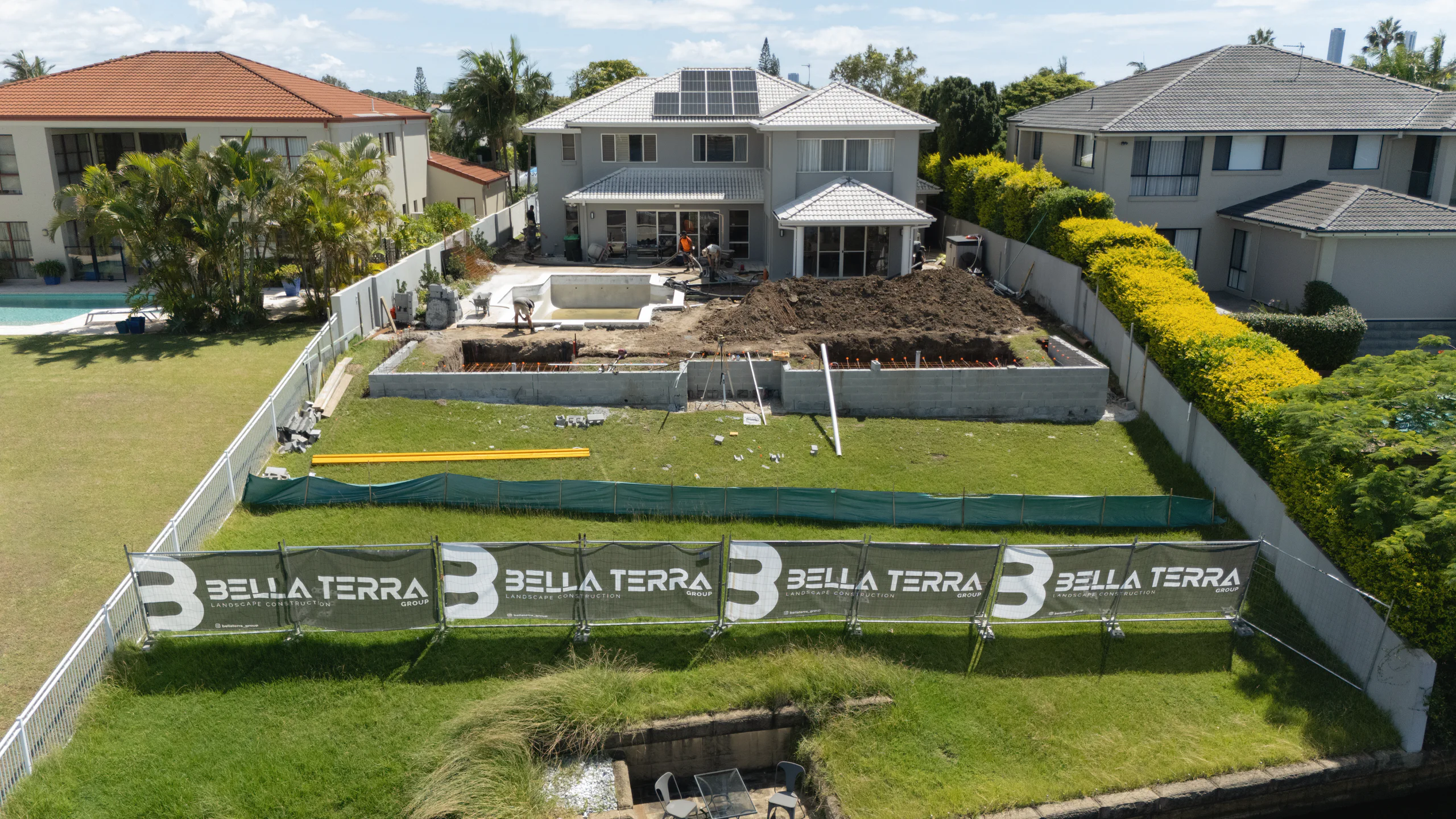 Drone view of a waterfront backyard mid-build: new block walls, the pool shell and the dig, behind Bella Terra site banners