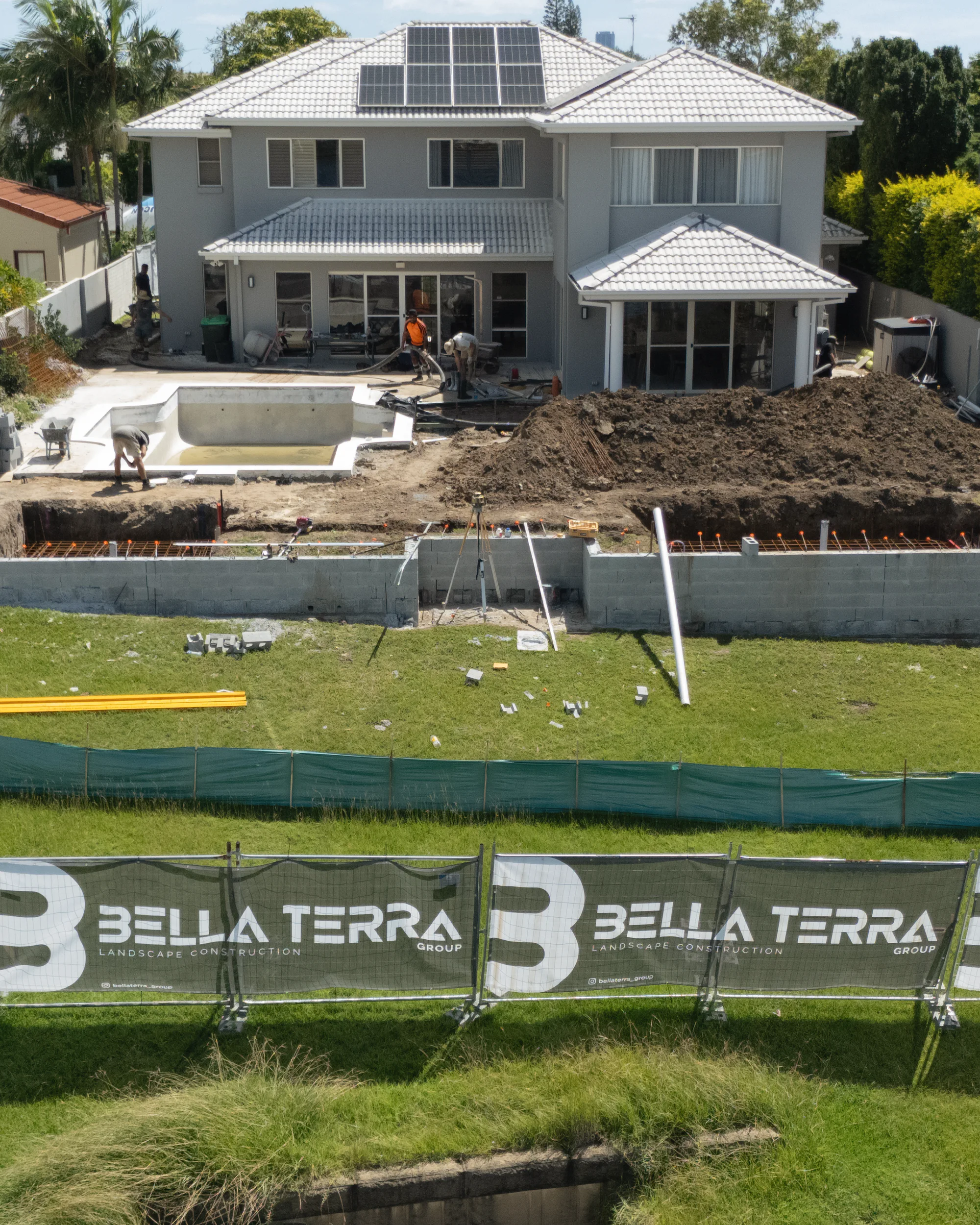 Drone view of a waterfront backyard mid-build: new block walls, the pool shell and the dig, behind Bella Terra site banners